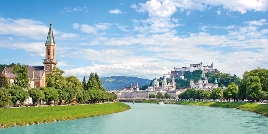 The Salzburg skyline on a bright day: a red brick church, buildings with round copper roofs and the bright blue river water can be seen. 